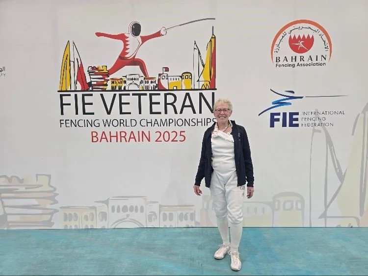 Sylvia Brown in fencing attire standing in front of a large wall mural with the dae and mural for the FIE Veteran Fencing World Championships