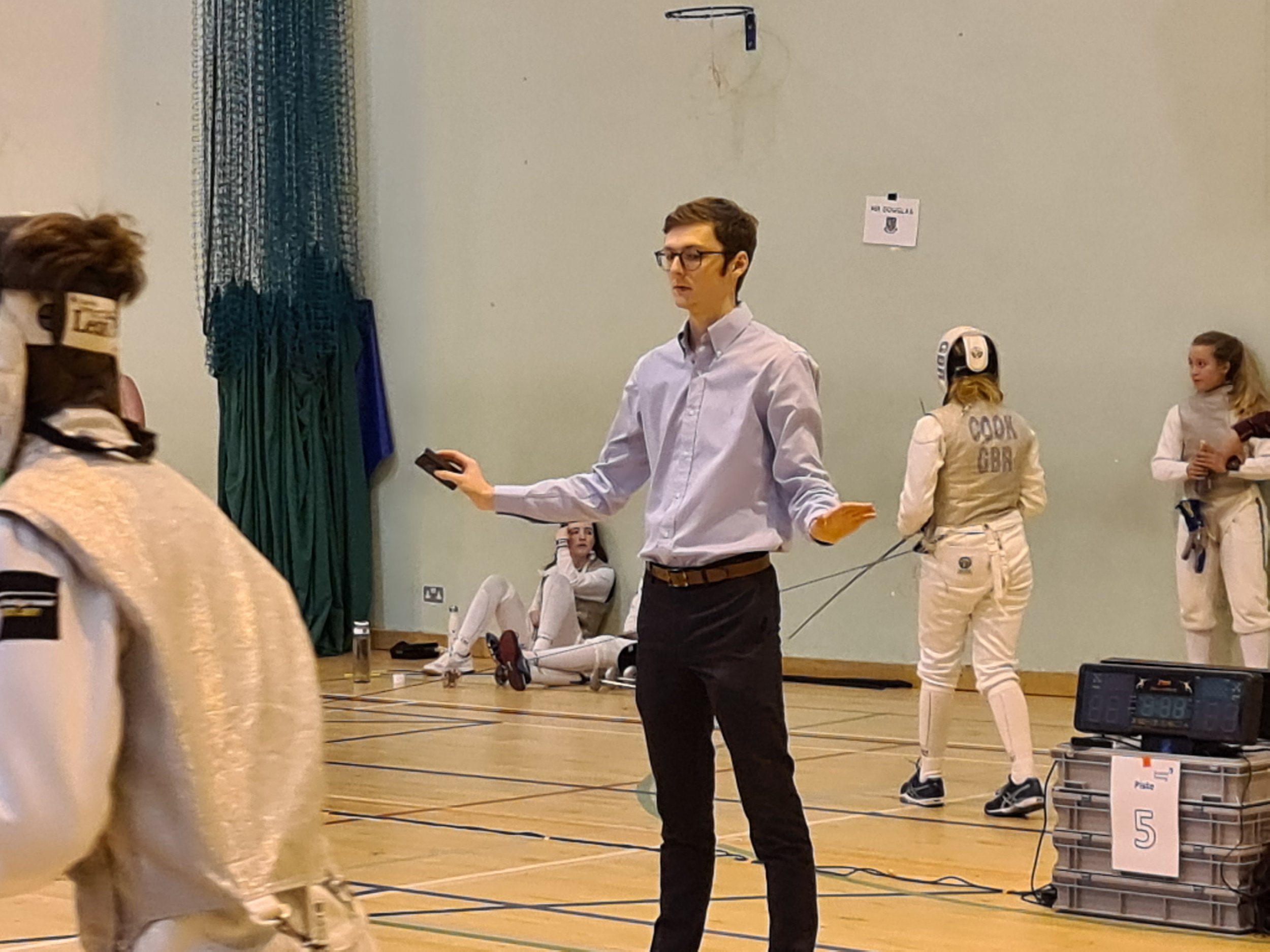 Picture of a referee in the engaurde hand signals with some fencers in the background